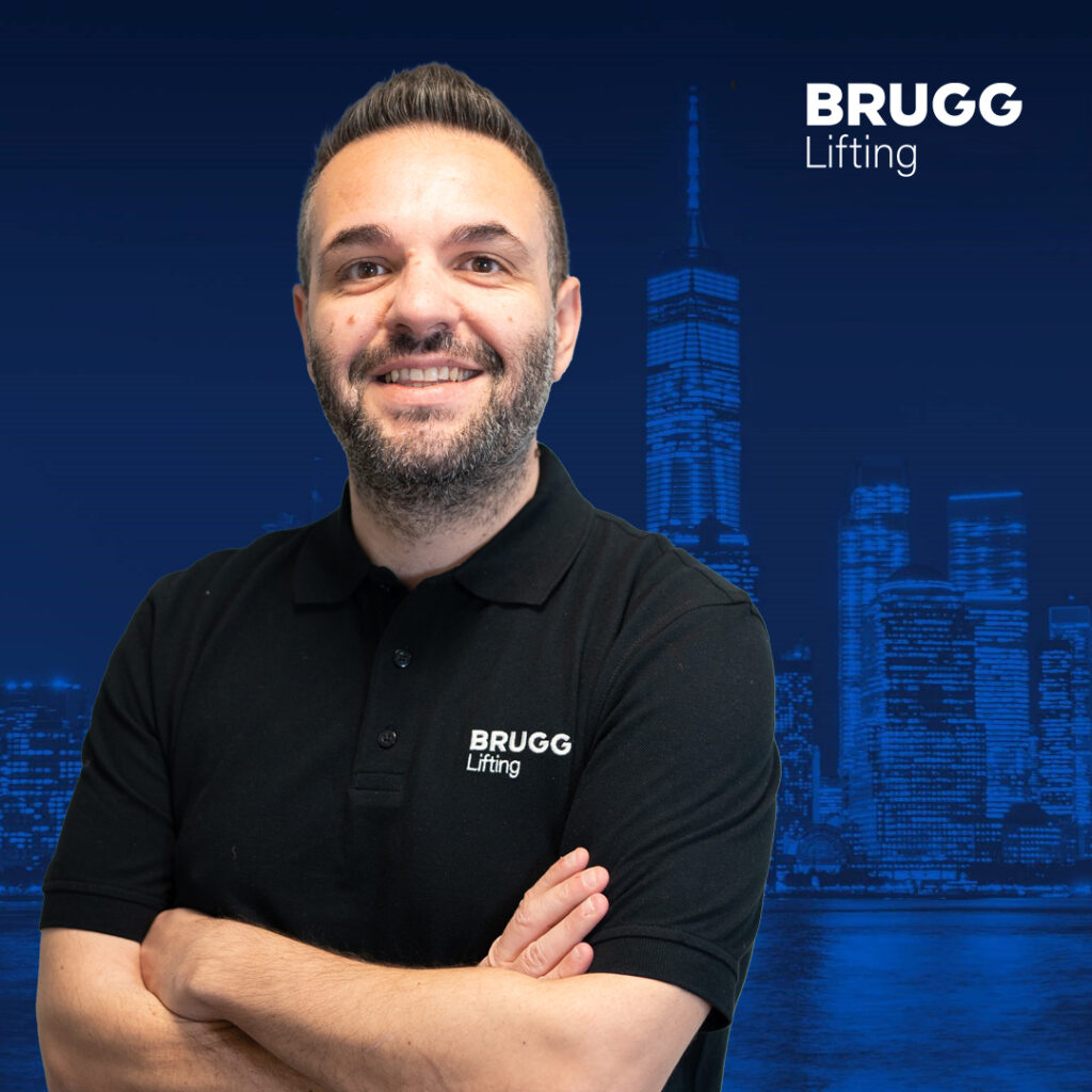 Christos Tsinalidis, Mitarbeiter von Brugg, lächelt seitlich mit verschränkten Armen in die Kamera. Er trägt ein schwarzes BRUGG Polo-Shirt und steht vor einem blauen Hintergrund.